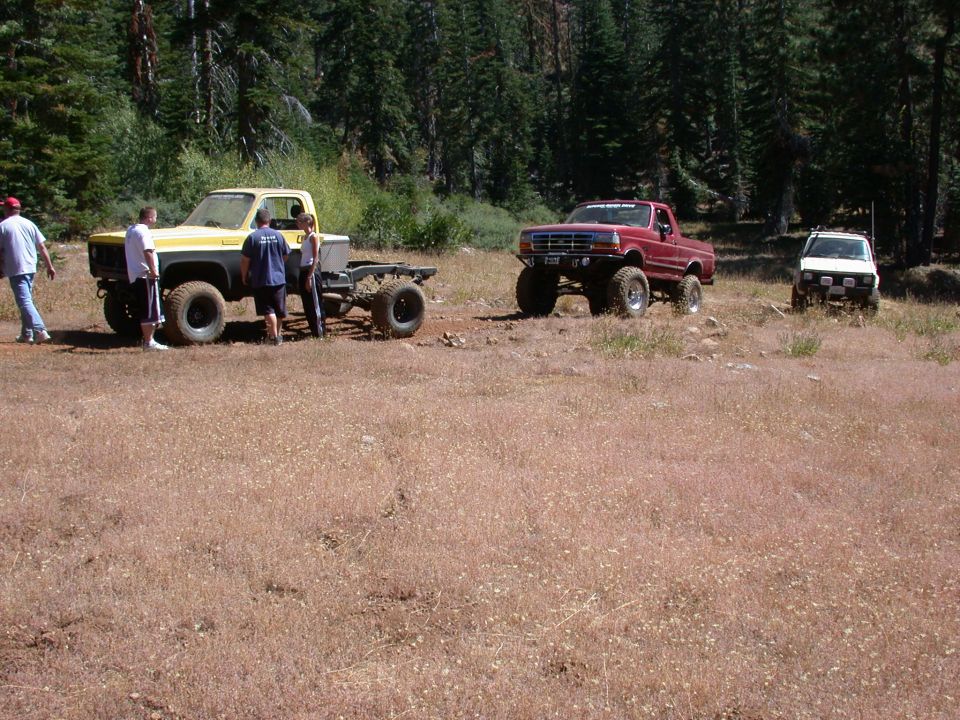 Yellow and grey pickup truck chassis with large tires, parked in a grassy field with trees