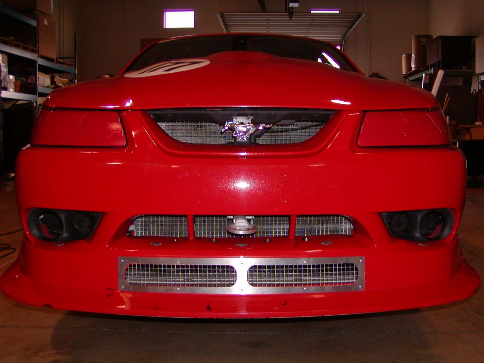 Front view of red Ford Mustang race car with mesh grille, tow hook, and number roundel on hood in a garage