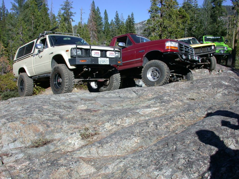 Three modified off-road trucks parked on a rocky incline with trees in the background
