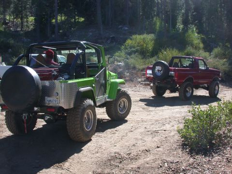 1996 Ford Bronco - Two modified off-road vehicles are parked on a dirt path surrounded by trees.