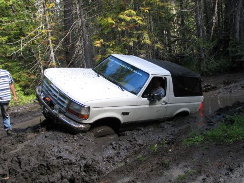 1995 Ford Bronco - A white 1995 Ford Bronco is stuck in deep mud on a wooded trail, with a person observing from the side.