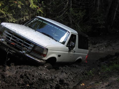 1995 Ford Bronco - A white 1995 Ford Bronco navigates a muddy off-road trail, its tires deep in the mire.