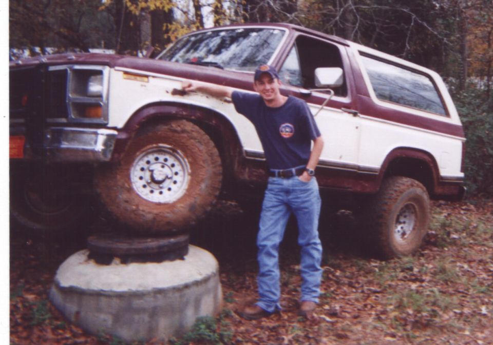 Man in blue shirt and jeans leans on a lifted 1980s Ford Bronco with two-tone paint
