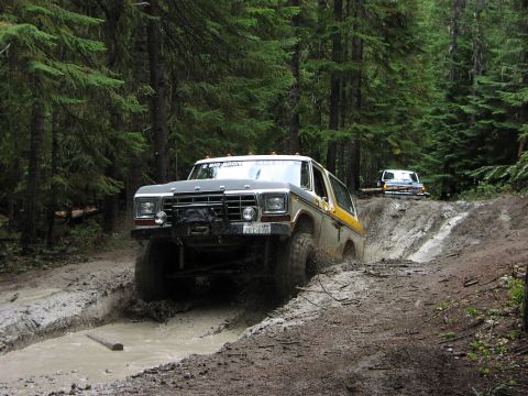 1995 Ford Bronco - Two Ford Broncos navigate a challenging muddy off-road trail in a forest.