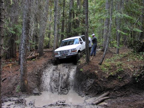 1995 Ford Bronco - A white 1995 Ford Bronco tackles a challenging muddy obstacle on a forest trail.