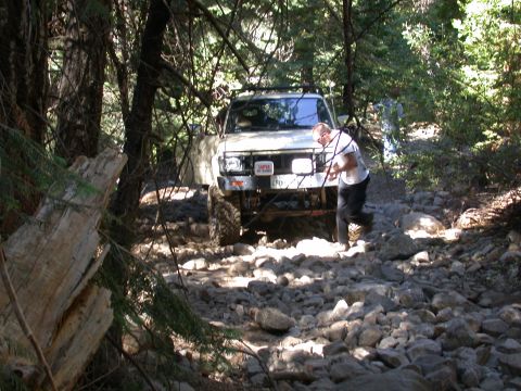 1996 Ford Bronco - A person stands by a white SUV as it drives over a rough, rocky path in a wooded area.