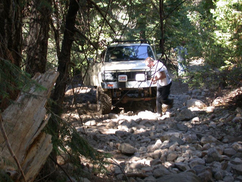 White SUV navigating a very rocky forest trail. A person is near the front of the vehicle