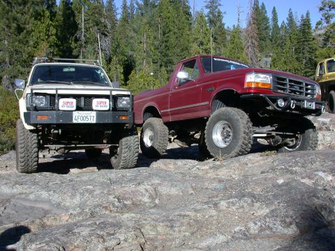 1996 Ford Bronco - Two modified trucks, a 1996 Ford Bronco and a white SUV, are parked on a rocky incline.