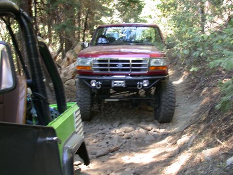 1996 Ford Bronco - Two off-road vehicles navigate a rocky, wooded trail. The front vehicle is a red Ford truck.