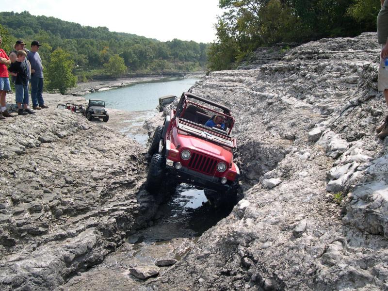 Red open-top Jeep Wrangler TJ rock-crawling through a limestone gully beside a river as spectators watch