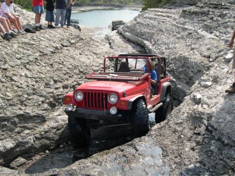 1999 Jeep TJ - A red Jeep Wrangler drives through a rocky, water-filled ravine with spectators watching from above.