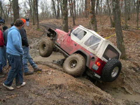 1999 Jeep TJ - A red and white 1999 Jeep TJ navigates a challenging muddy trail with onlookers watching.