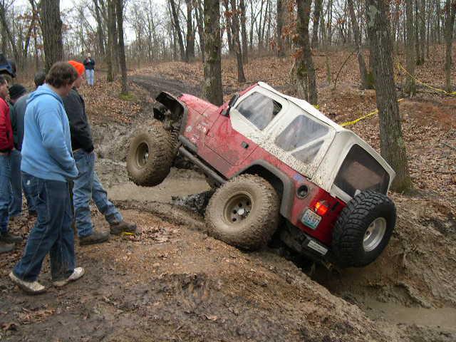 Muddy red Jeep Wrangler on big tires climbing a steep rut on a wooded off-road trail with spectators.