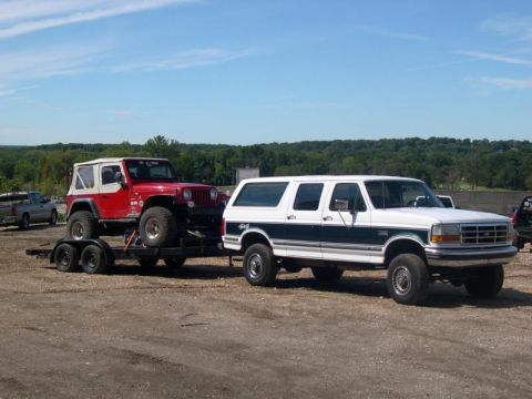 1992 Ford C-250 (Centurion Conversions) - White Ford SUV towing a red Jeep Wrangler on a trailer under a blue sky.