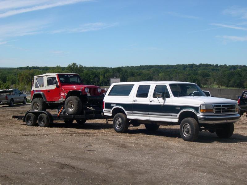 White/green Ford Centurion SUV towing trailer with red Jeep Wrangler on gravel lot