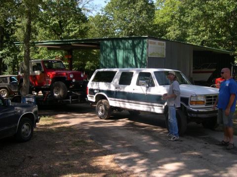 1992 Ford C-250 (Centurion Conversions) - A white and teal SUV is parked on a dirt lot next to two men, with a red Jeep on a trailer in the background.