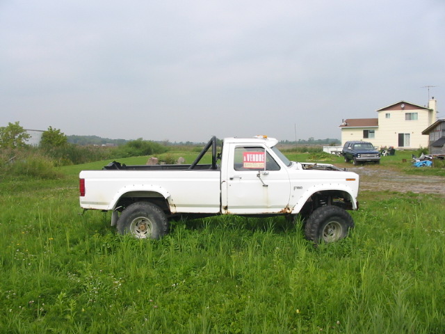 White pickup truck with off-road tires and roll bar, parked in grass. Another dark SUV is visible in the background near a house