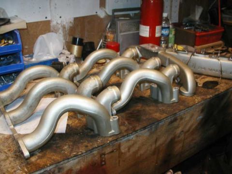1993 Ford Taurus - Several silver exhaust manifolds are laid out on a cluttered wooden workbench.