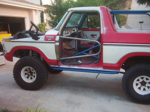 1978 Ford Bronco - Driver-side profile of a red and white lifted Ford Bronco parked in a residential driveway with its doors, top and interior trim removed, e…