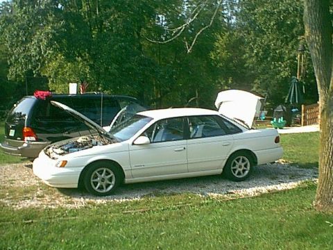 1994 Ford Taurus - A white sedan sits in a yard with its hood and trunk open, parked beside a dark minivan.