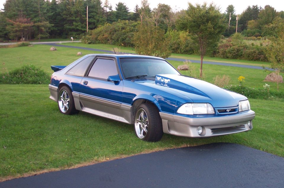 Blue and silver Fox-body Mustang GT hatchback on grass with chrome wheels and hood scoop