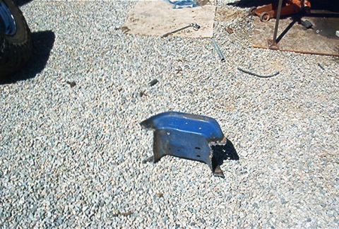 1986 Ford Bronco - A blue metal bracket with mounting tabs sits on a gravel surface.