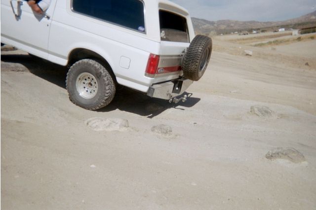 Rear of a white Ford Bronco with spare tire flexing over sandy terrain
