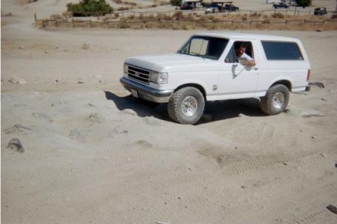 1990 Ford Bronco - lifted white Ford Bronco sits on a sandy off-road course as the driver leans out the window, RVs in the distance.