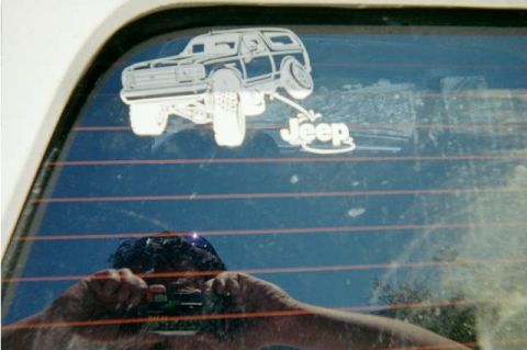 1990 Ford Bronco - white SUV-shaped sticker beside Jeep lettering on a defrost-line rear window, with the photographer reflected holding a camera.
