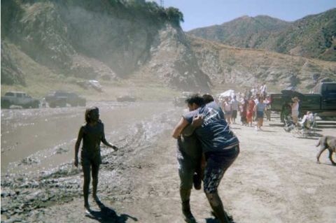 1990 Ford Bronco - Mud-covered people walk ashore from a muddy wash as a crowd, trucks and a dog gather along the bank in a dusty canyon.