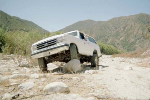 1990 Ford Bronco - silver Ford Bronco lifts its front wheel onto a rock in a dry riverbed surrounded by green mountains.