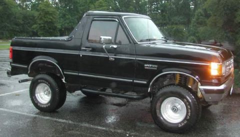 1988 Ford Bronco - A black Ford Bronco is parked on a wet surface, showing its front three-quarter view with aftermarket side steps.