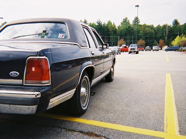 Rear view of a dark blue 1990 Ford Crown Victoria parked in a lot. Other cars are visible in the background
