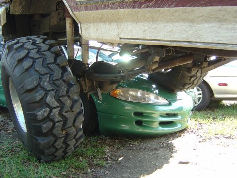 1993 Ford Bronco - A lifted truck's massive tire rests on the hood of a green Dodge Avenger, crushing its front end.