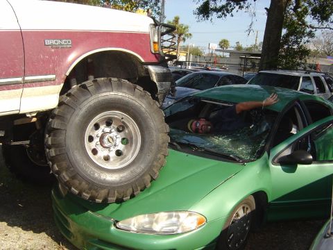 1993 Ford Bronco - A lifted Ford Bronco appears to have driven over a green sedan, causing significant damage to the car's roof and windshield.