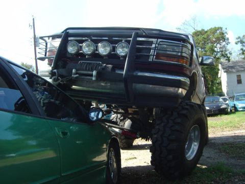 1993 Ford Bronco - A heavily modified truck is positioned over a green car, with other vehicles visible in the background.