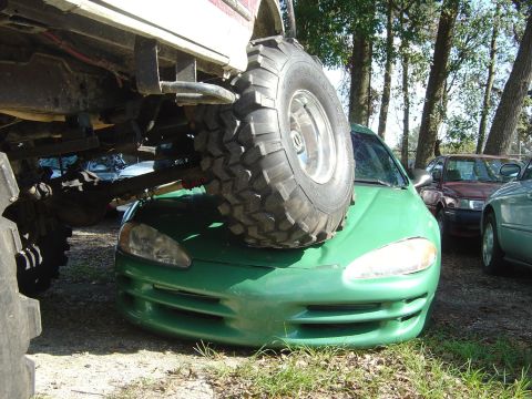 1993 Ford Bronco - A large off-road tire rests on the hood of a green car, with other vehicles visible in the background.