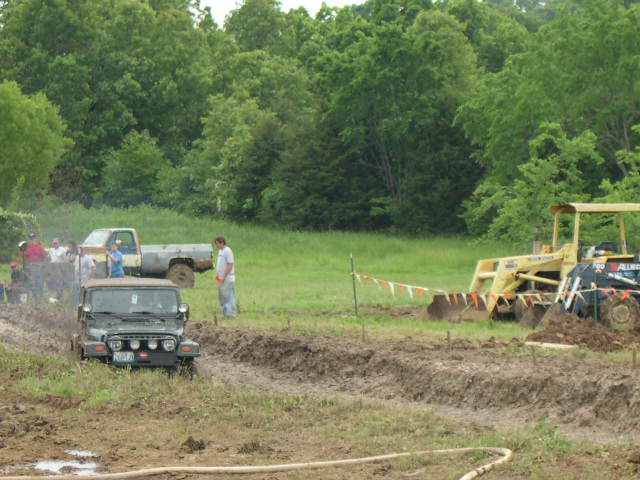 Dark green Jeep Wrangler TJ driving through mud course with tractor and pickup nearby