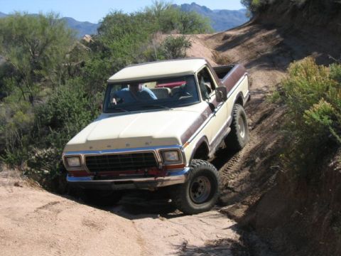 1979 Ford Bronco - Three-quarter front view of a topless cream-over-maroon Ford Bronco descending a steep dirt trail cut into a desert hillside, with two occu…