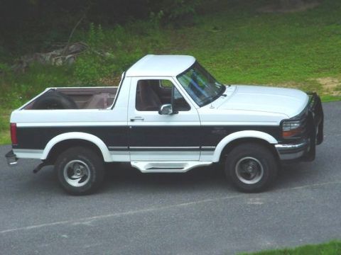 1979 Ford Bronco - Passenger-side profile of a white-over-dark-blue two-tone Ford Bronco with the rear hardtop removed, parked on pavement beside a grassy ban…