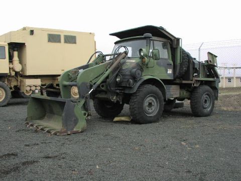 1981 Ford Bronco - A military front loader with a large bucket is parked on gravel, with another military vehicle to its left.