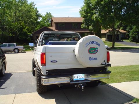1993 Ford Bronco - This white Ford Bronco is parked in a driveway, showing its rear end with a custom spare tire cover.