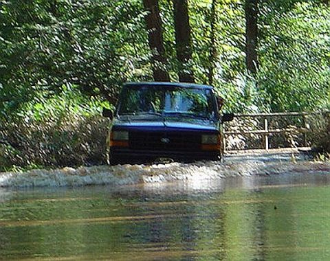 1989 Ford Ranger - A blue 1989 Ford Ranger navigates through a flooded area, with trees and a wooden fence visible in the background.