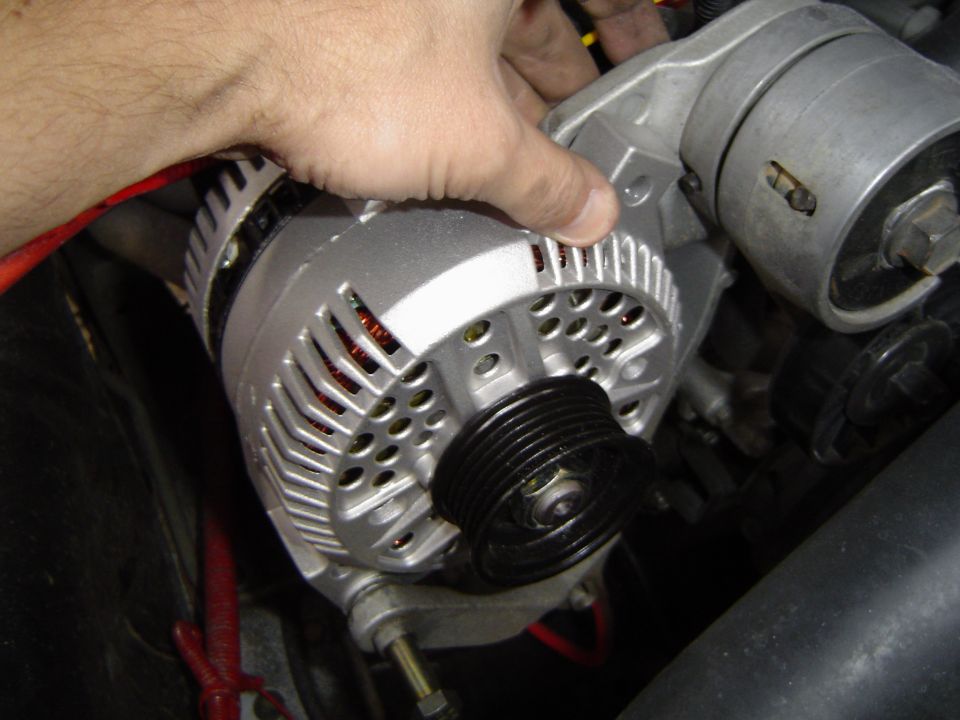 Hand holding a freshly rebuilt silver alternator in an engine bay beside red wiring