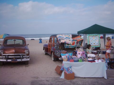 1978 Ford Bronco - Two classic cars are parked on the sand near a beach vendor's stall under a green canopy.