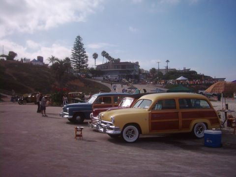 1978 Ford Bronco - Several classic woodie wagons are lined up on a paved area with a beach and buildings in the background under a partly cloudy sky.