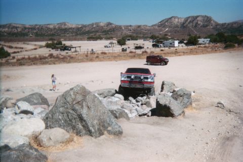 1990 Ford Bronco - red truck climbs rocks rear-on at a desert OHV staging area as a child walks nearby and RVs sit in the distance.