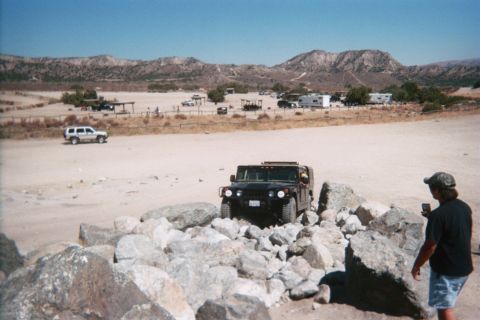 1990 Ford Bronco - black Hummer crawls a rock pile while a man watches and SUVs sit across a desert staging area with RVs beyond.