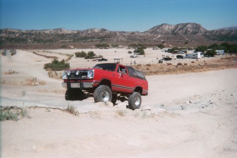 1990 Ford Bronco - lifted red soft-top 4x4 descends a white sandy trail with a desert campground and mountains in the distance.