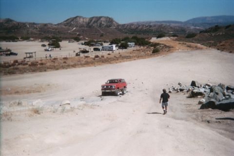 1990 Ford Bronco - red SUV is parked on a sandy slope while a man walks a trail in a desert OHV area dotted with RVs and rocks.